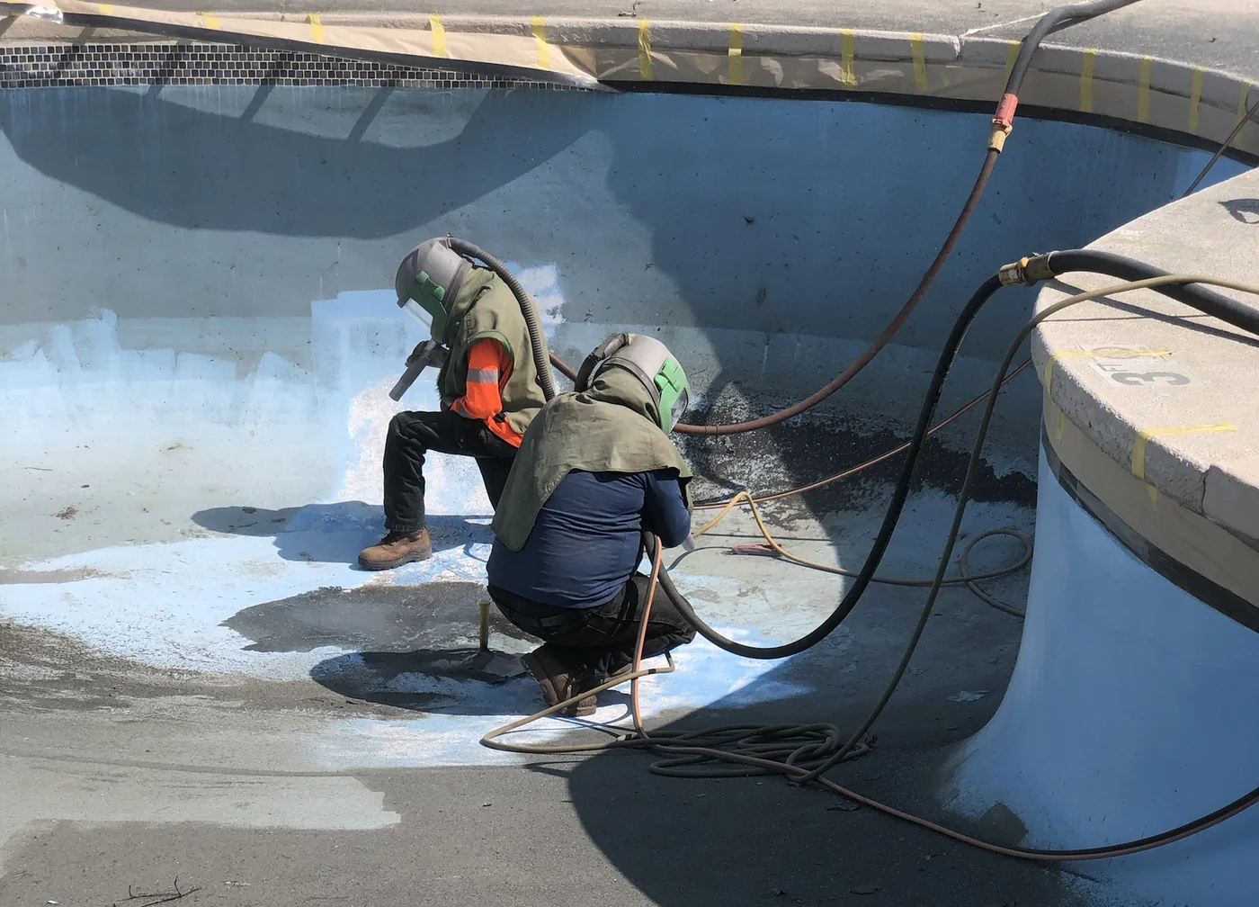 Two of the crew in blast hoods stripping a pool on site, hoses run out across the deck